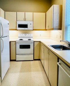 Kitchen with white appliances including refrigerator, hood microwave, oven, and sink in front of window; cream colored cabinets and flooring